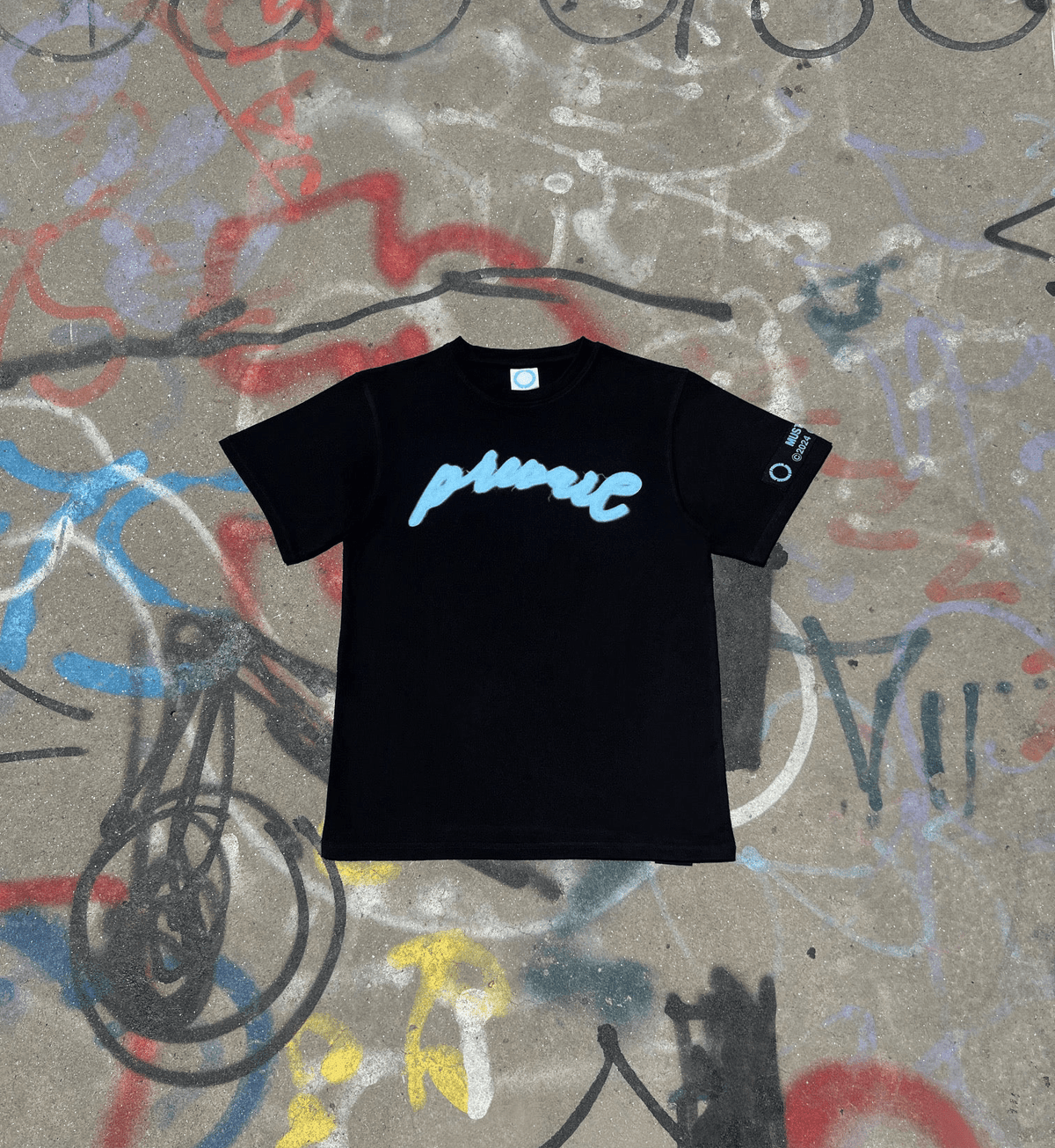 A flat lay of the MUST PREVAIL black statement tee on a graffiti-covered concrete surface. This shot features the teal blue 'Prevail' DTG graphic and highlights the 425 GSM heavyweight cotton texture, thick ribbed collar, and oversized boxy fit. The industrial background reinforces the gritty, urban architectural design of the garment.