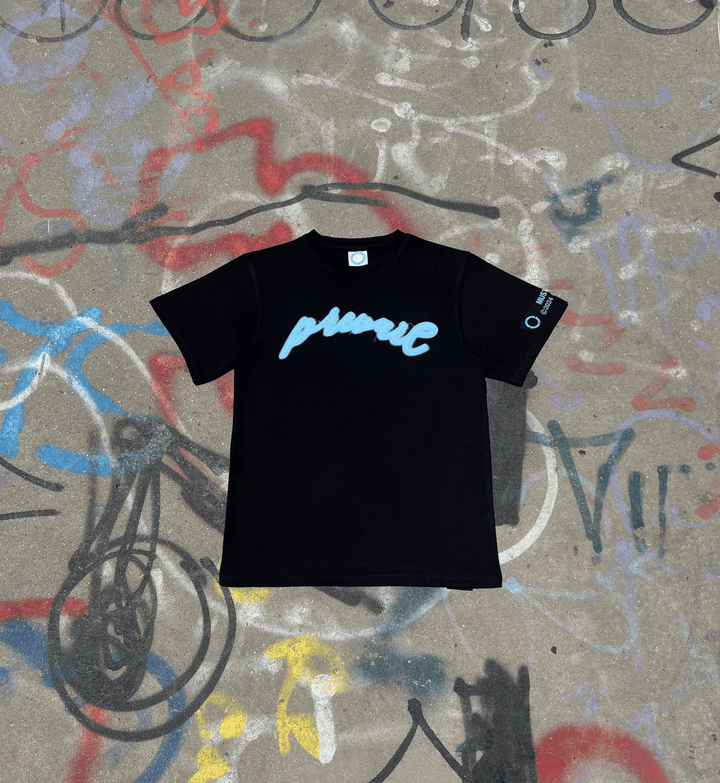 A flat lay of the MUST PREVAIL black statement tee on a graffiti-covered concrete surface. This shot features the teal blue 'Prevail' DTG graphic and highlights the 425 GSM heavyweight cotton texture, thick ribbed collar, and oversized boxy fit. The industrial background reinforces the gritty, urban architectural design of the garment.