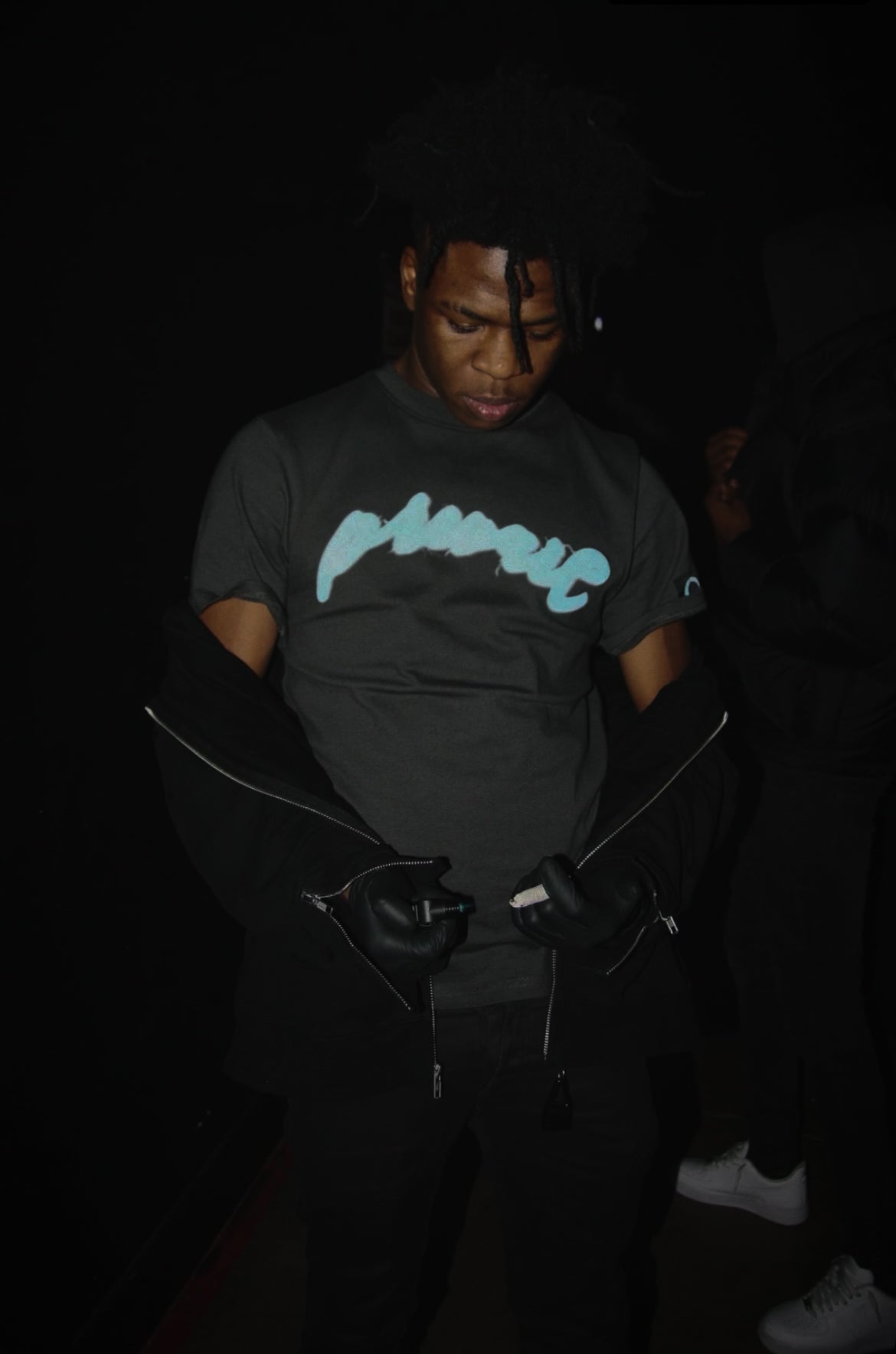 A flash-photography night lifestyle shot of a model wearing the MUST PREVAIL grey heavyweight cotton statement tee. Features the blue 'Prevail' DTG graphic, styled with black leather gloves and dark layering. The image highlights the structured boxy silhouette and oversized fit in an urban outdoor setting at night.