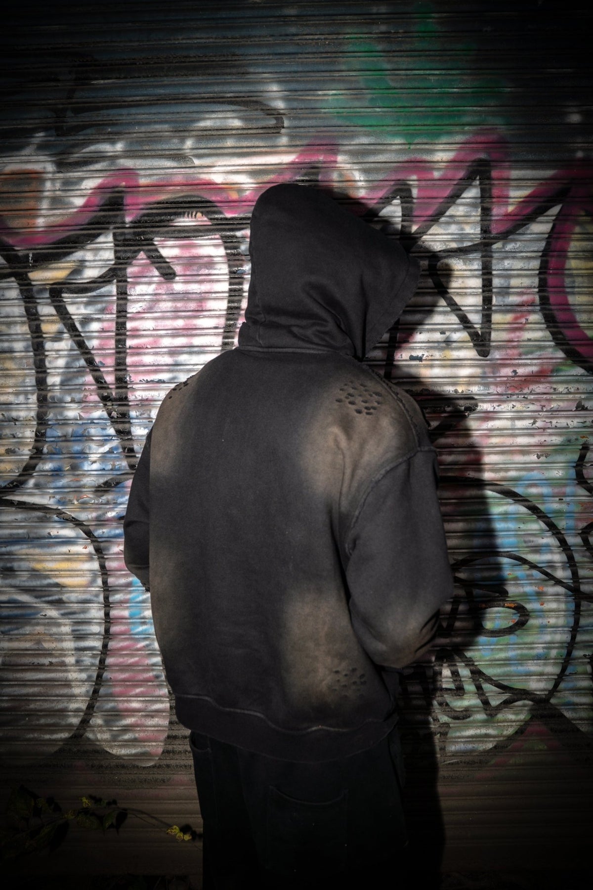 A back view lifestyle shot of the MUST PREVAIL Y2K heavyweight zip-up hoodie. This image highlights the technical sun-faded wash and custom distressing on the shoulders and back. The model is standing against a graffiti-covered industrial roll-up door, showcasing the oversized boxy silhouette and premium fleece texture.