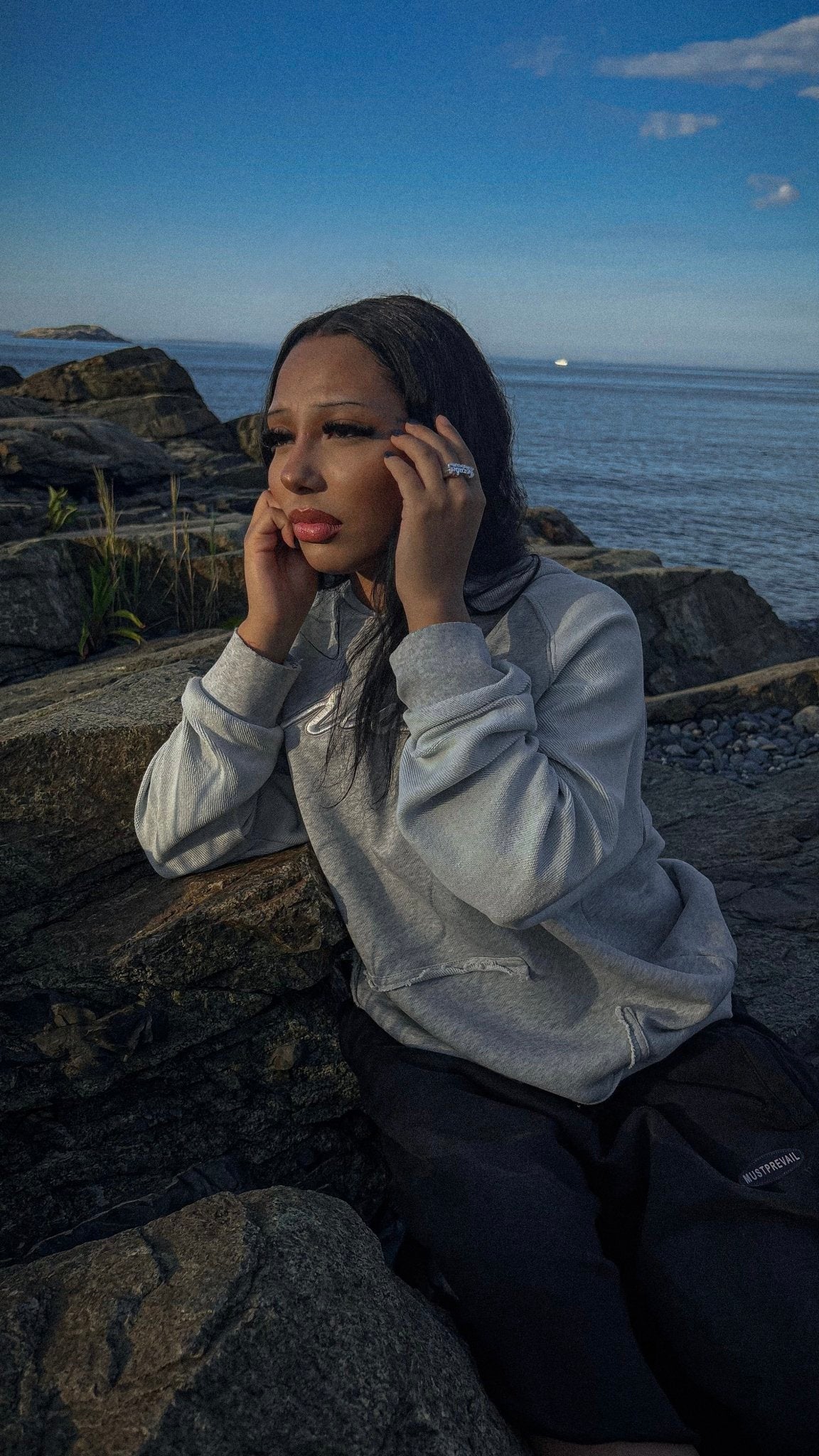 An on-body candid shot of a 5’7 female model in front of the rocks on the beachside shoreline in Nahant, Massachusetts. Featuring the “emo” hoodie complimented by a saddled raglan-sleeve with drop shoulders, silk-threaded embroidery, French Terry paneling and a Heathered grey torso. Details include a distressed/frayed “ghost” kangaroo pocket, silk-thread torso embroidery, two-tone sleeve paneling, a saddled-raglan sleeve, and screen-printed silk woven tags