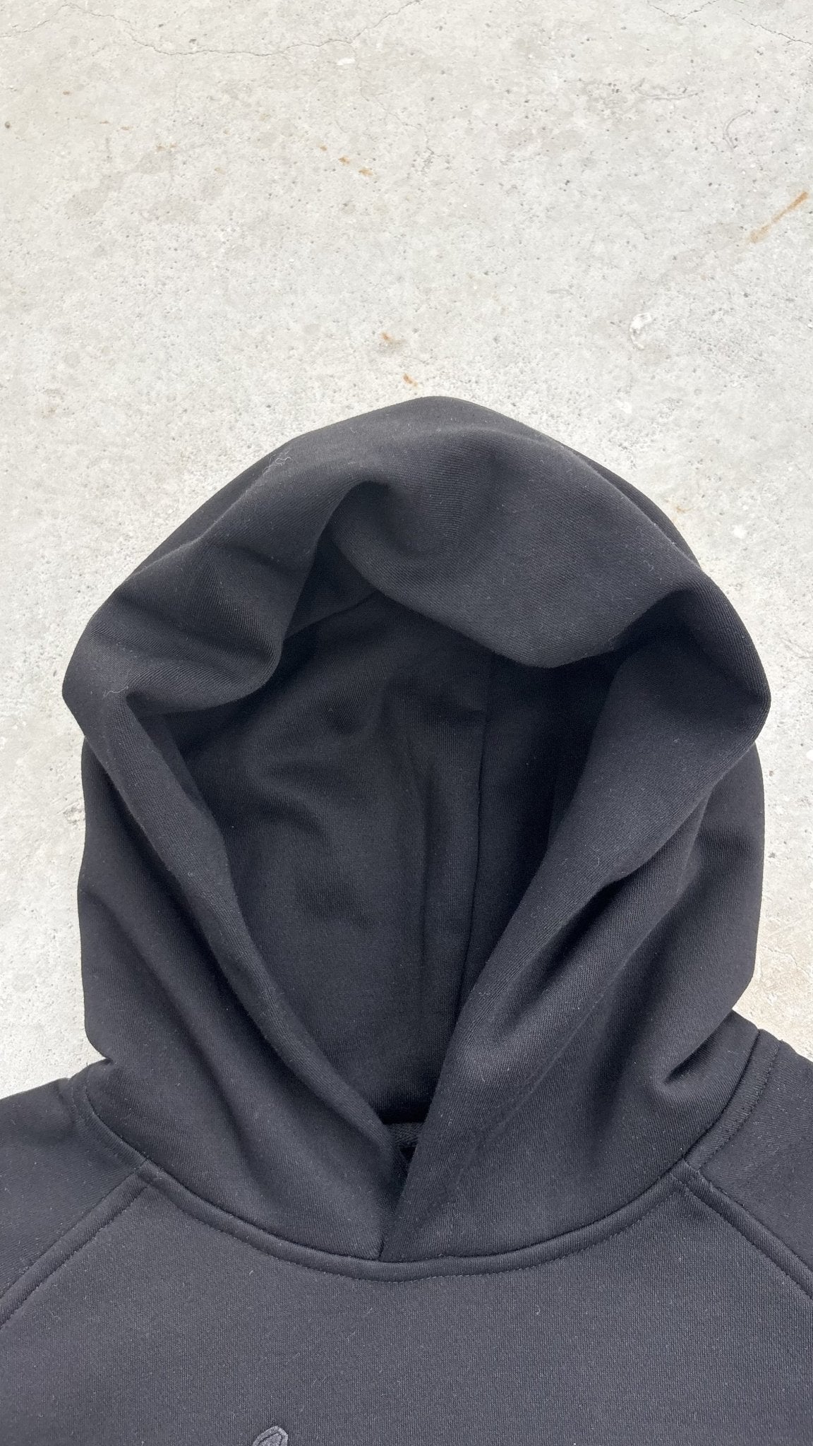 A top-down macro flat lay shot focused strictly on the hood structure of the black Onyx Hoodie, with the hood occupying 50 percent of the frame against a concrete garage floor. The close-up layout profiling highlights the 420 GSM heavyweight cotton-blend fabric density, thick double-layered hood construction, clean seam finishing, and the custom woven "V₂" neck tag visible at the collar line.