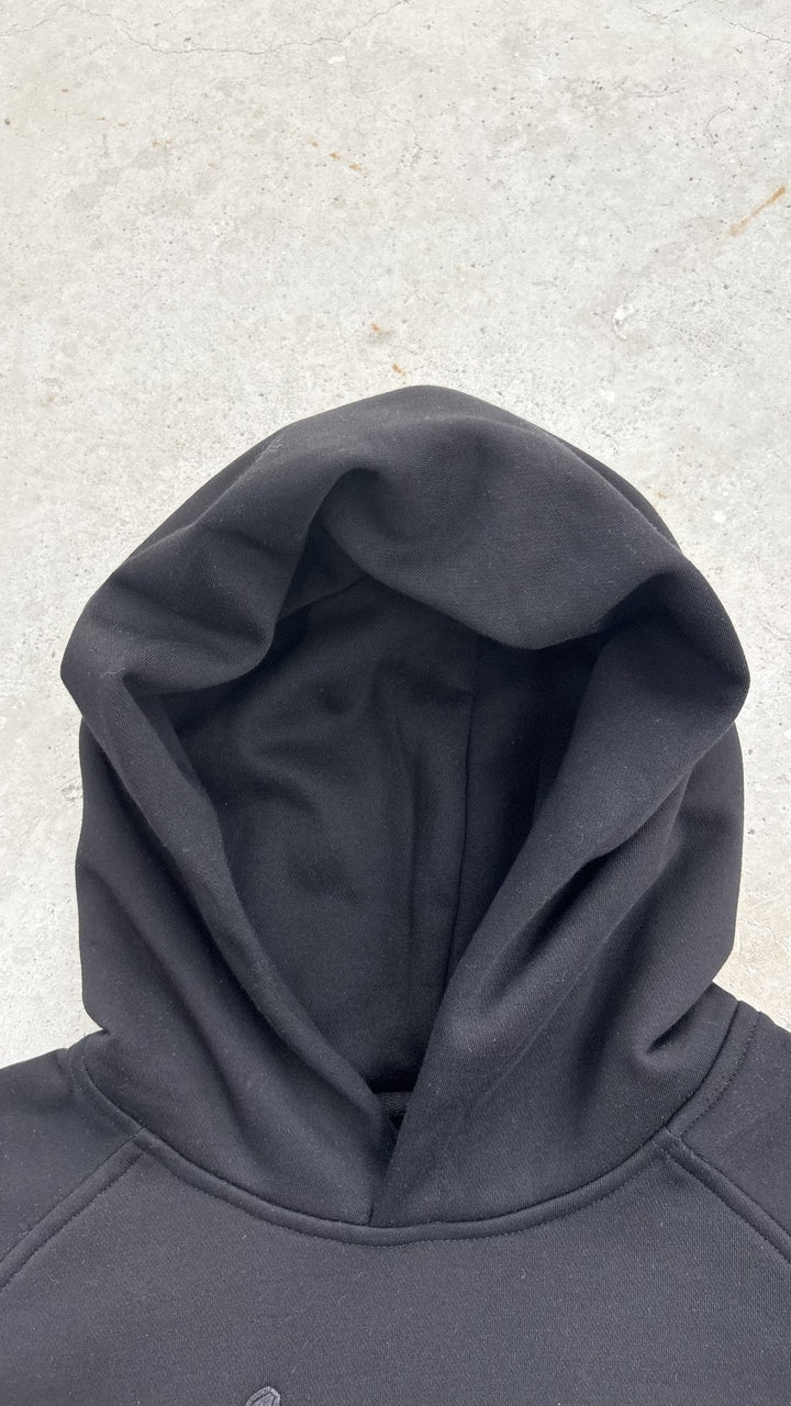 A top-down macro flat lay shot focused strictly on the hood structure of the black Onyx Hoodie, with the hood occupying 50 percent of the frame against a concrete garage floor. The close-up layout profiling highlights the 420 GSM heavyweight cotton-blend fabric density, thick double-layered hood construction, clean seam finishing, and the custom woven "V₂" neck tag visible at the collar line.