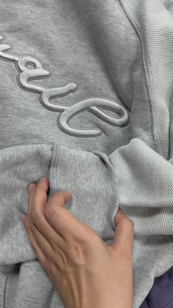 A macro video frame close-up focusing on the dynamic texture of the Emo Hoodie. The close-up shot glides slowly over the 420 GSM heavyweight French Terry cotton fabric, capturing the contrasting two-tone paneling, the fine sheen of the silk-threaded tonal torso embroidery, and the detailed raw loops of the interior fleece structure as the light moves across the garment.