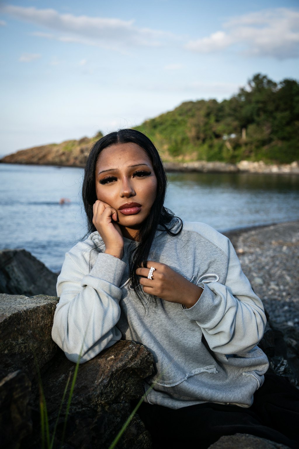 A straightforward on-body editorial shot of a 5’7 female model facing directly forward with no hand raised, positioned in front of the coastal rocks along the beachside shoreline in Nahant, Massachusetts, wearing the Emo Hoodie. The centered angle emphasizes the 420 GSM heavyweight French Terry cotton construction featuring a heathered grey torso, two-tone sleeve paneling, and saddled-raglan drop-shoulder structure. Key details highlight the distressed and frayed deconstructed "ghost" kangaroo pocket, silk-