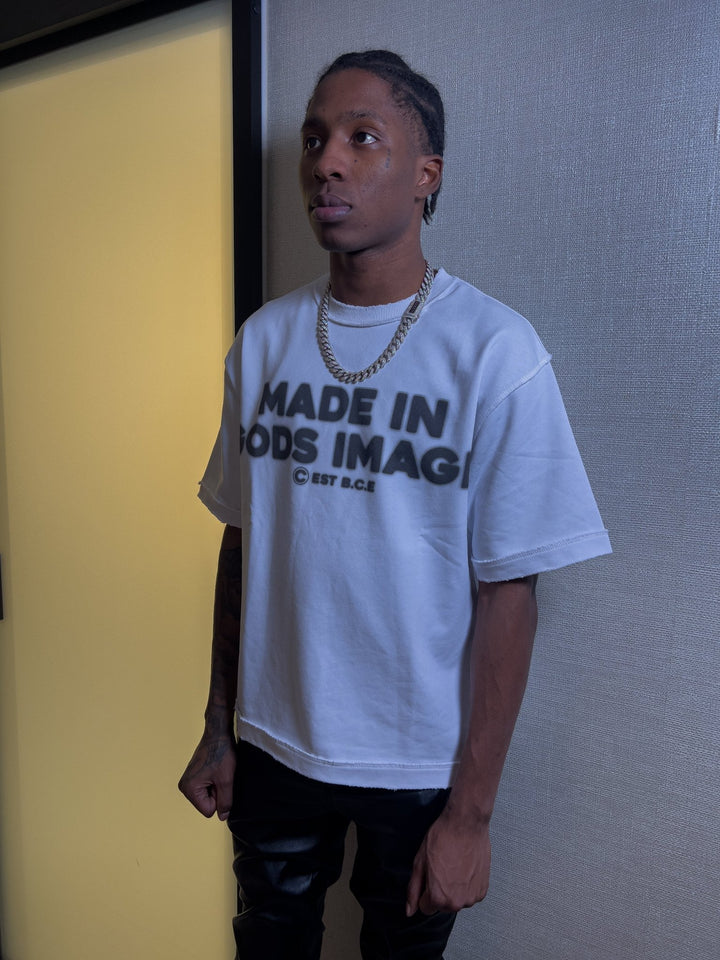 A front-facing studio portrait of a model wearing the MUST PREVAIL white 425 GSM heavyweight French Terry inside-out tee. The image features high-contrast lighting that highlights the technical inside-out stitching, distressed hems, and DTG chest print. Styled with a gold chain against a plain background to showcase the oversized boxy silhouette.