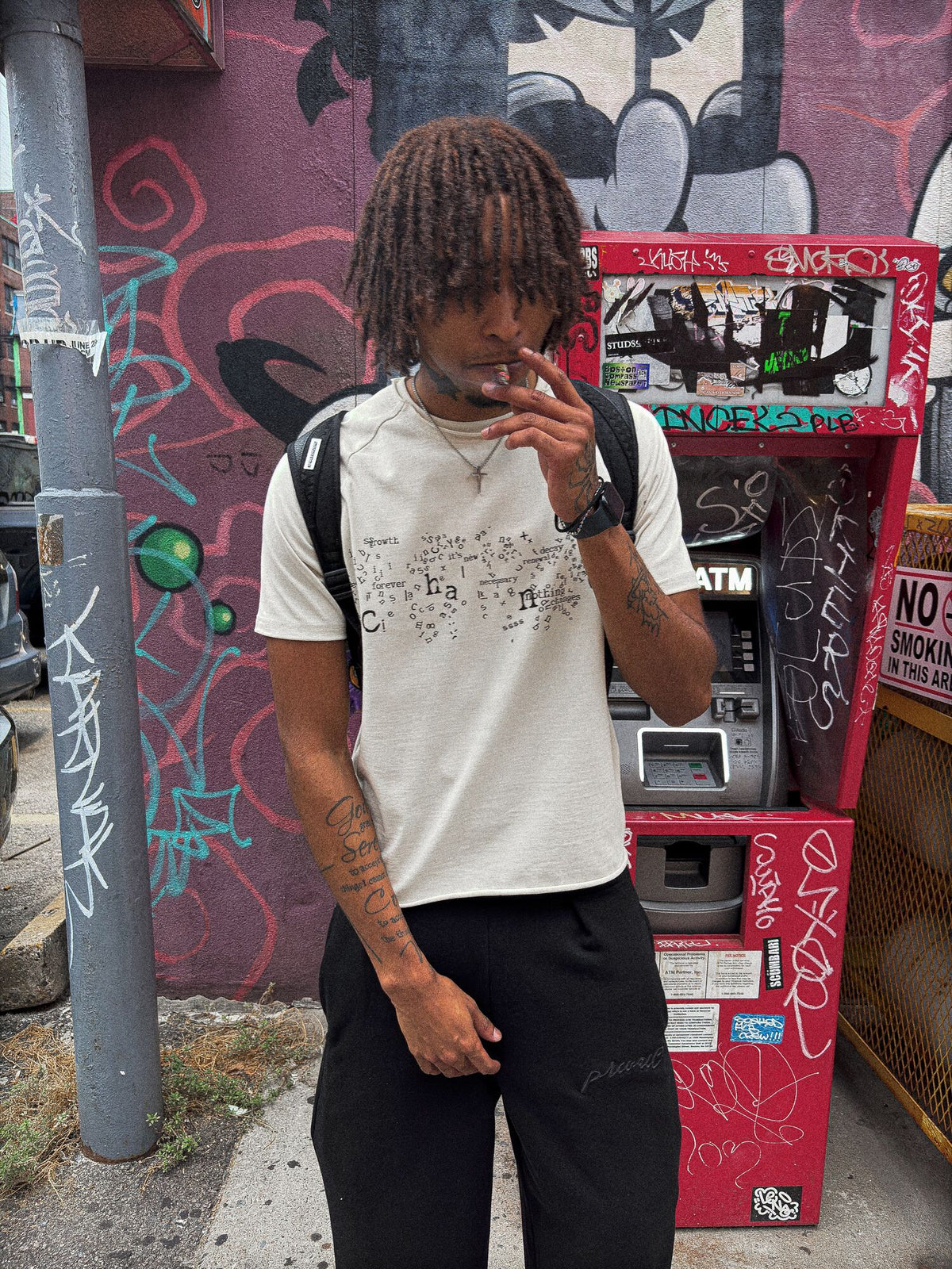 A zoomed-out 70-degree angled candid shot of a 6’1 light-skinned model with butterscotch-complexioned skin and reddish amber-brown dreads, captured smoking in front of the Chinatown gate ATM in Boston while wearing the "poem" tee paired with the Onyx Sweats. The dynamic wide perspective highlights the 325 GSM French Terry fleece-cotton tee, complimented by saddled-raglan sleeve paneling with a slim drop-shoulder fit, a direct-to-garment torso print, a stacked raw-edge ribbed neckline, and hand-cut Dremel mi